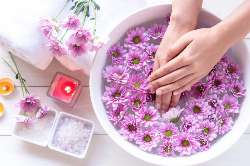 A person soaks their hands in a white bowl filled with water and floating pink daisies. Nearby are candles, bath salts, a towel, and more flowers, creating a relaxing spa atmosphere.