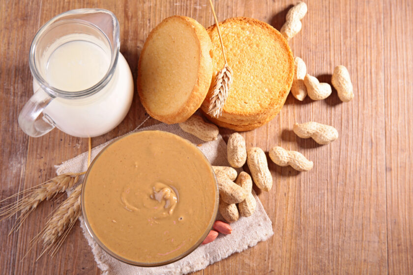 A bowl of peanut butter, a glass jug of milk, round cookies, and scattered peanuts on a wooden table with wheat stalks.