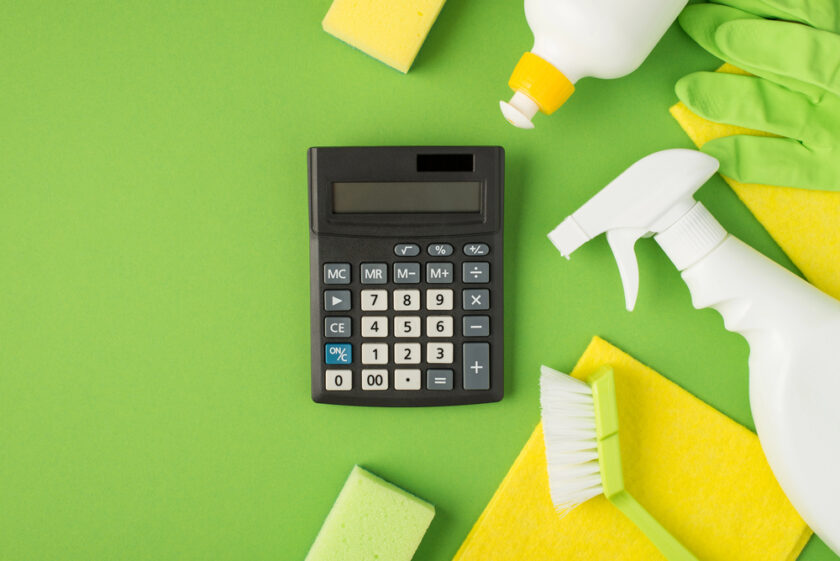 A black calculator lies on a green surface surrounded by cleaning items, including a spray bottle, gloves, sponges, a scrubbing brush, a yellow cloth, and a bottle of cleaning liquid.