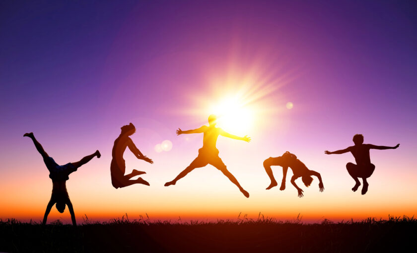 Five people are silhouetted against a vibrant sunset sky, jumping and doing acrobatic poses in mid-air on grassy ground, with the sun shining brightly in the background.