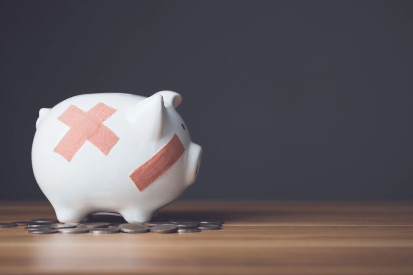 A white piggy bank with two adhesive bandages on its side sits on a wooden surface surrounded by scattered coins, against a dark gray background.