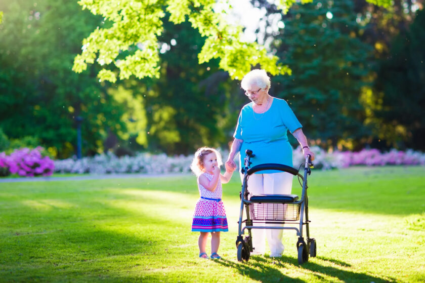 An elderly woman with a walker holds hands with a young girl in a colorful dress. They are smiling and walking together in a sunlit park with green grass and trees in the background.