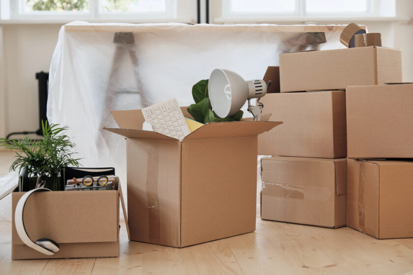 Cardboard moving boxes, some open and some closed, are scattered on a wooden floor. Items like a lamp, keyboard, plant, and headphones are visible, with a covered piece of furniture in the background.
