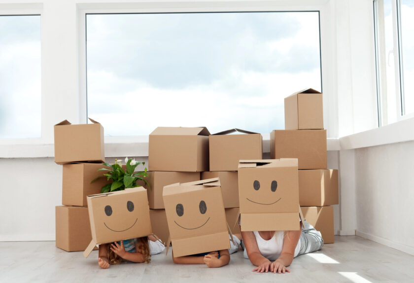Three people lying on the floor with cardboard boxes drawn with smiley faces on their heads, surrounded by more moving boxes and a potted plant in a bright room with large windows.