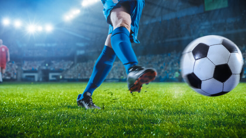 A soccer player in blue uniform kicks a soccer ball on a brightly lit stadium field, with a blurred crowd and another player visible in the background.