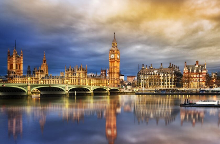 The Palace of Westminster and Big Ben are illuminated at sunset, reflecting on the River Thames in London, with dramatic clouds in the sky.