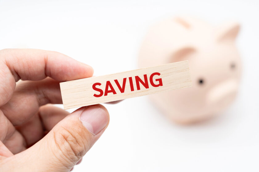 A hand holding a wooden block with the word SAVING in red letters, with a blurred pink piggy bank in the background.