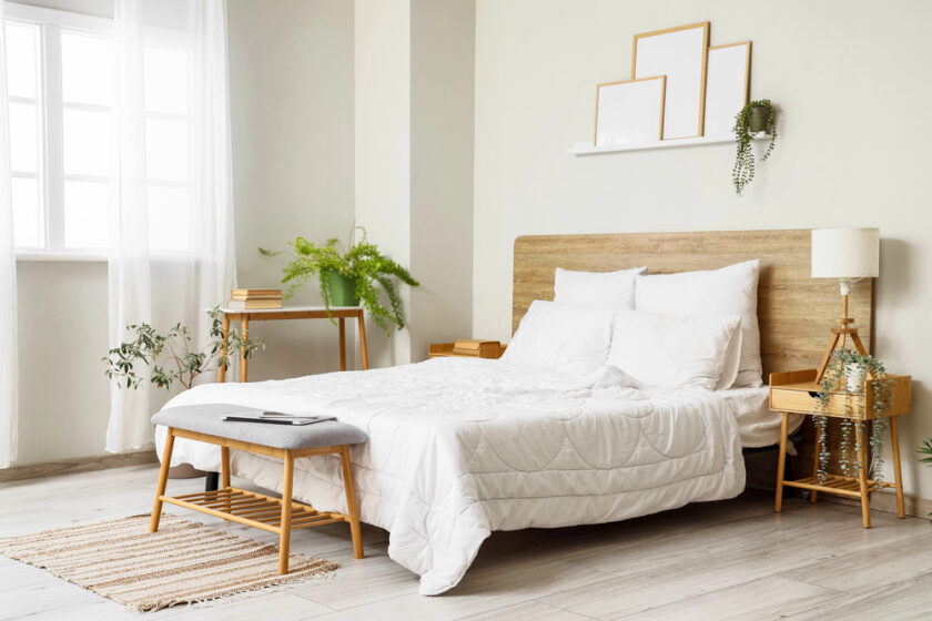 A bright, modern bedroom with a wooden bed frame, white bedding, and pillows. There are plants, books, a lamp, framed art on the wall, and sunlight streaming through sheer curtains over a large window.