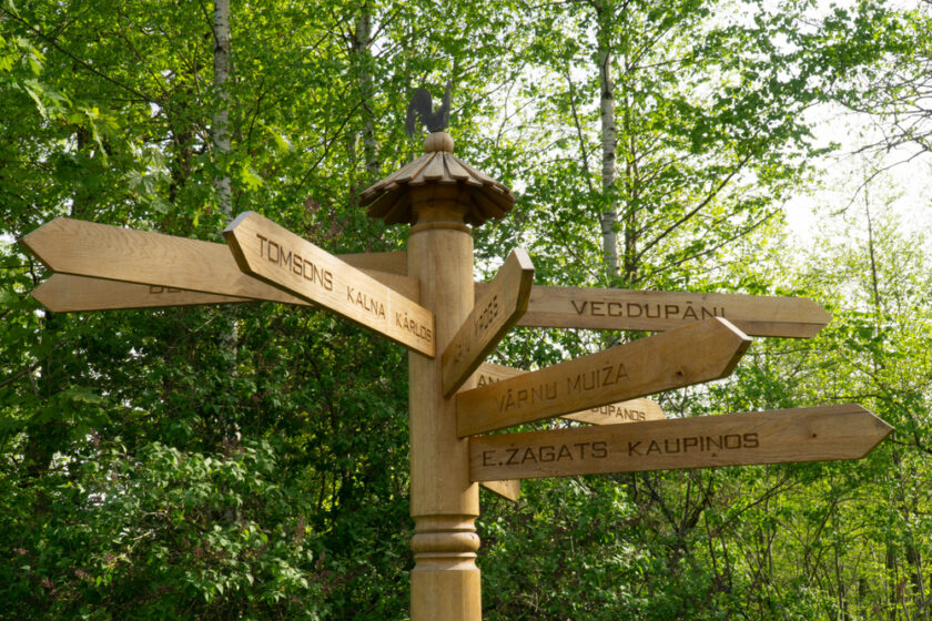A wooden signpost with multiple directional arrows displaying Latvian place names, standing in a green, leafy forested area on a sunny day.