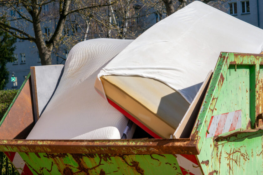 Two old mattresses are placed inside a large, green metal dumpster outdoors, with trees and an apartment building visible in the background.