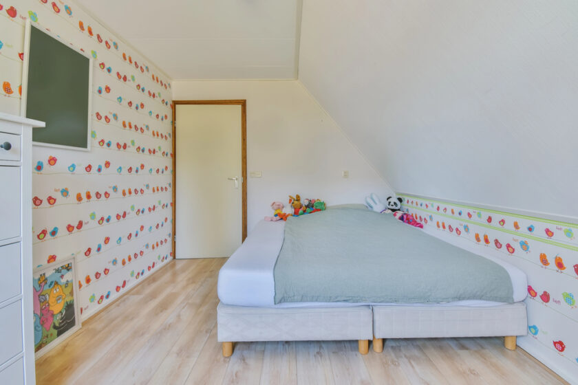 A bright childrens bedroom with colorful cartoon wallpaper, a neatly made bed with a light blue duvet, stuffed animals on the bed, wood flooring, a flat-screen TV on the wall, and a closed door.