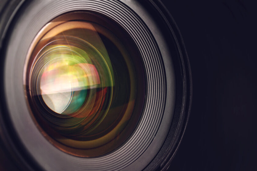 Close-up of a camera lens showing reflections, colorful light flares, and detailed concentric rings around the glass elements against a dark background.