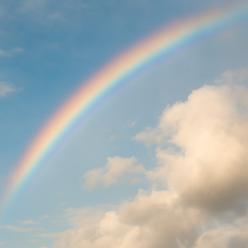 A bright, colorful rainbow arcs across a partly cloudy sky, with soft white clouds and a blue background.