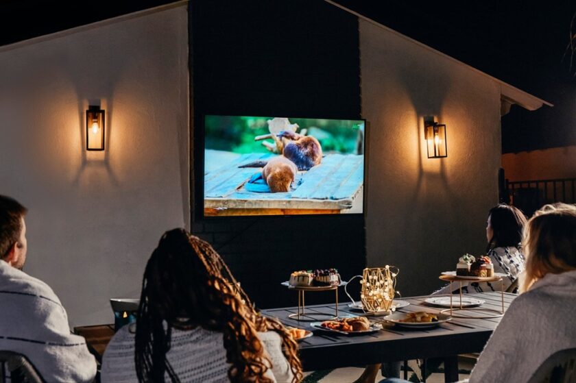 Four people sit outside at night watching a nature show on a wall-mounted TV, with food and desserts on a table in front of them. The area is lit by wall lamps.