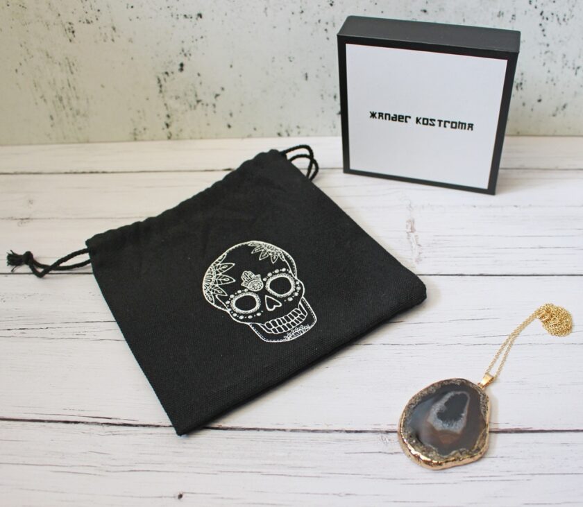 A black drawstring pouch with a white skull design, a slice of agate pendant on a gold chain, and a white box with black trim and text, all arranged on a white wooden surface.