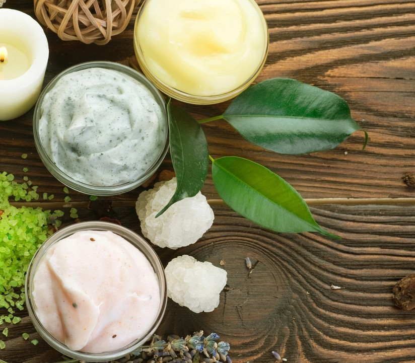 Three small glass jars of colorful creams or masks are arranged on a rustic wooden surface, surrounded by green leaves, bath salts, and decorative items, creating a natural spa-like setting.