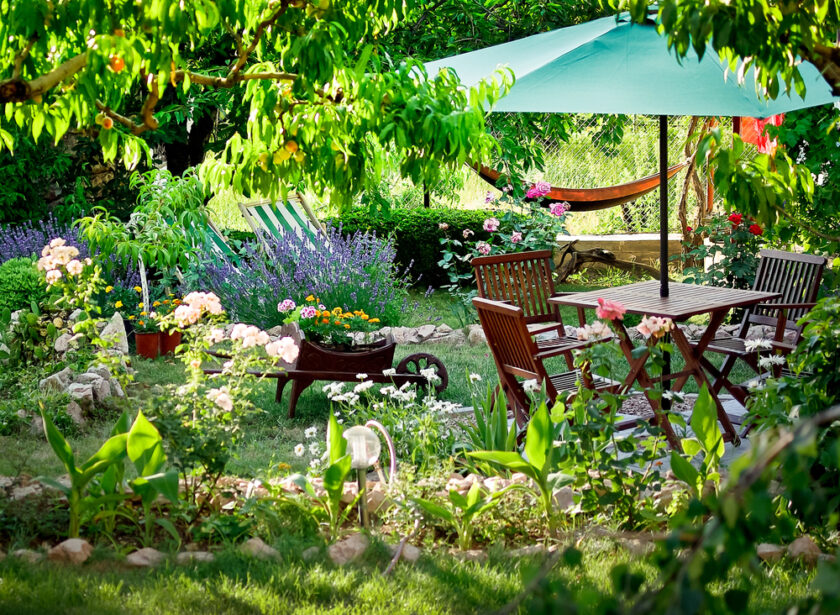 A lush garden with blooming flowers, green plants, a wooden table and chairs under a large green umbrella, a hammock in the background, and a wheelbarrow filled with flowers. Sunlight filters through the trees.