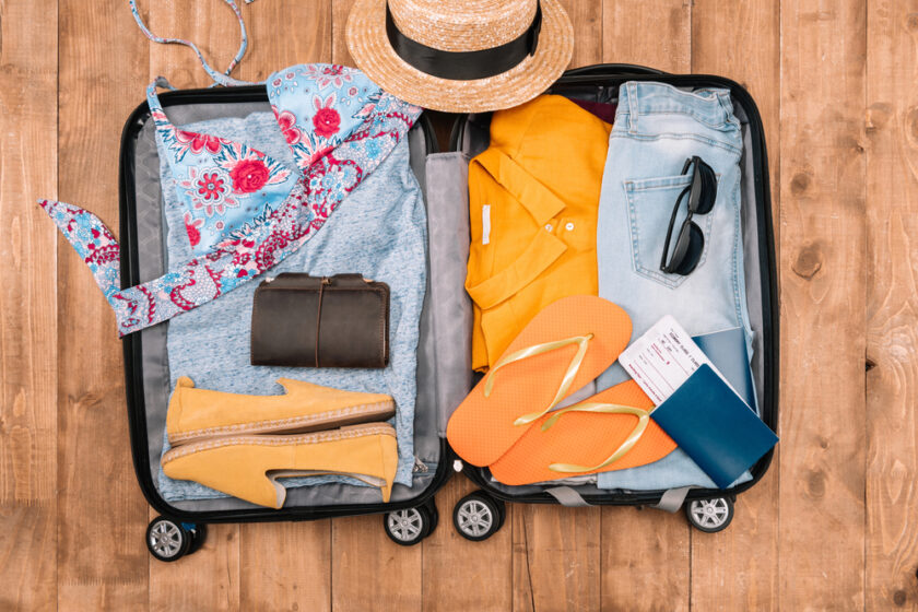 Open suitcase on a wooden floor packed with clothes, a floral bikini, yellow shirt, jeans, sandals, hat, sunglasses, a wallet, and travel documents including a passport and boarding pass.