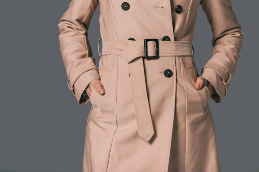 A person wearing a beige trench coat with black buttons and a belt stands with their hands in the coat pockets against a plain gray background.
