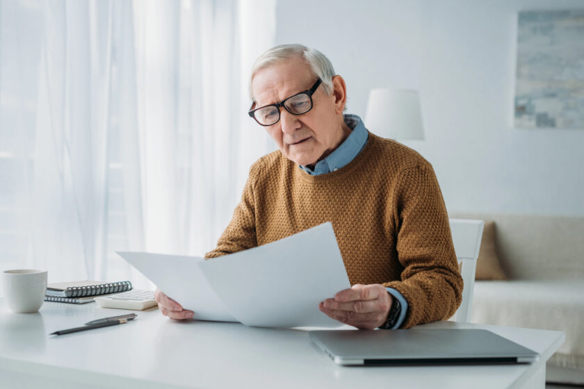 An older man with glasses sits at a white desk, reading documents. He wears a brown sweater and is surrounded by a laptop, notebook, pen, and coffee cup in a bright, modern room.