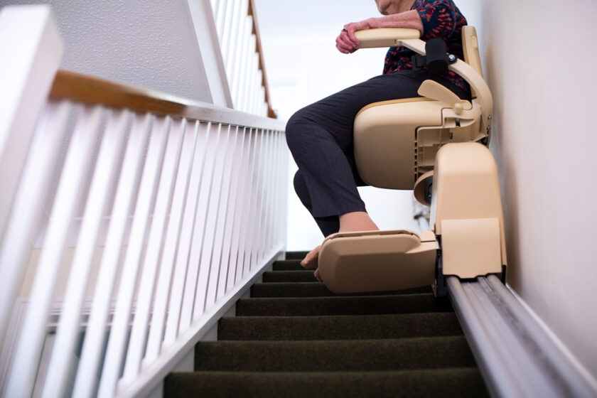 An older adult rides a stairlift chair up a carpeted staircase, with hands resting on the armrest and bare feet on the footplate, next to a white railing.