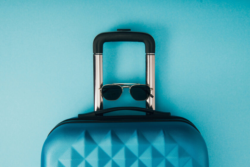 A blue suitcase with a textured surface stands upright against a light blue background. Sunglasses are hanging on the suitcase handle, creating a playful, travel-themed composition.