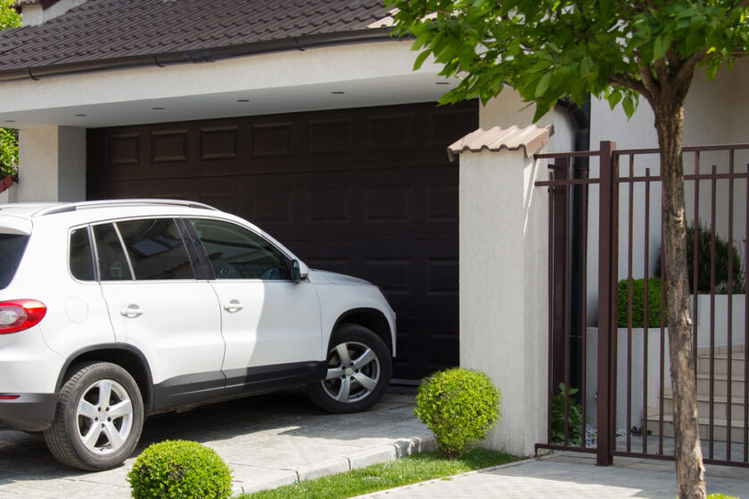 A white SUV is parked very close to a closed dark brown garage door in a driveway next to a modern house with a tree and trimmed bushes in the foreground.