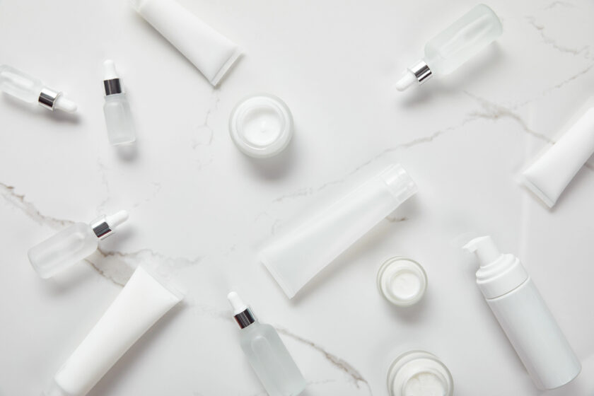 Several white and frosted glass skincare bottles, jars, and tubes are arranged on a white marble surface, viewed from above. The containers are minimalistic and unlabelled.