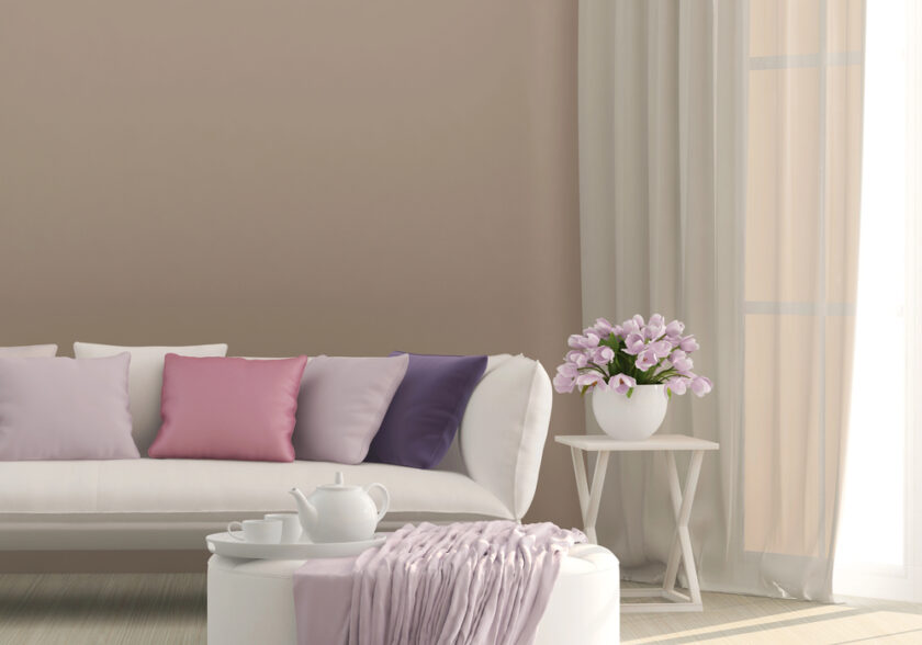 A modern living room with a white sofa, pastel pink and purple cushions, a round table with a teapot and cup, a pink blanket, a vase of pink flowers on a small side table, and light curtains by a window.