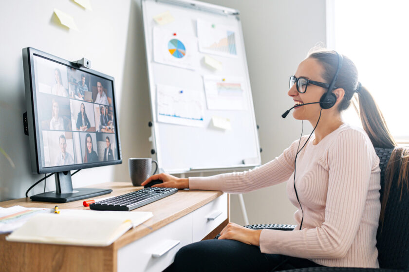 A woman wearing glasses and a headset sits at a desk, smiling at her computer screen during a video conference. A whiteboard with charts and sticky notes is in the background.