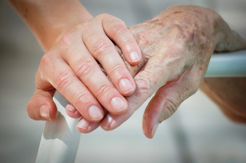 A young hand gently rests on top of an elderly hand holding a walker, symbolizing care, support, and intergenerational connection.