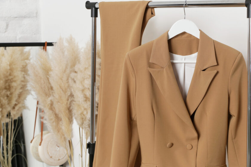 A tan blazer and matching pants hang on a clothing rack, with decorative pampas grass and a round woven bag in the background.