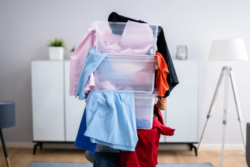 A person carrying several clear plastic storage bins overflowing with colorful clothes, obscuring their face, in a bright, modern room with white furniture and a standing lamp.