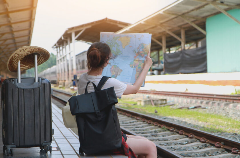 A woman with a backpack sits on a train platform reading a world map. A suitcase with a straw hat on top is beside her, and train tracks stretch into the distance under a partially covered station.