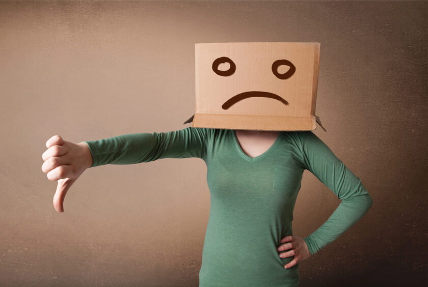A person wearing a green long-sleeve shirt with a cardboard box on their head showing a sad face. They are giving a thumbs-down gesture with their right hand.