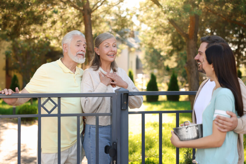 Two older adults smiling and greeting a younger couple at a gate outdoors. The younger woman is holding a pot, and they all appear happy and engaged in conversation with trees and greenery in the background.