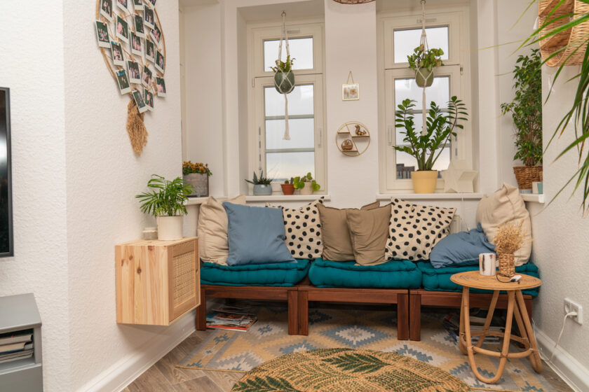 A cozy, light-filled nook with a teal-cushioned wooden bench, assorted pillows, potted plants, a small round table with a mug, and hanging photos on white walls beneath two windows.