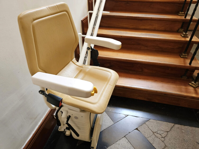 A beige stairlift chair with armrests is installed next to a wooden staircase, providing accessibility for individuals with mobility challenges. The chair is positioned at the bottom of the stairs.