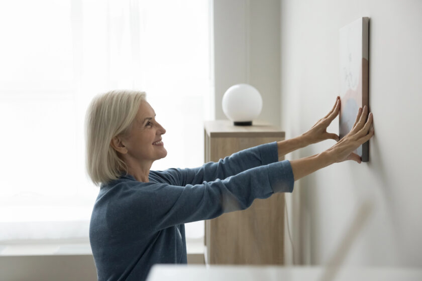A smiling older woman with short gray hair hangs a picture frame on a white wall in a bright, modern room with minimalistic decor. A spherical lamp sits on a wooden cabinet behind her.