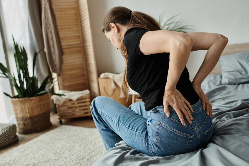 A woman in a black shirt and jeans sits on a bed, holding her lower back with both hands, appearing to be in pain. The room has a plant, a woven basket, and natural light coming in.