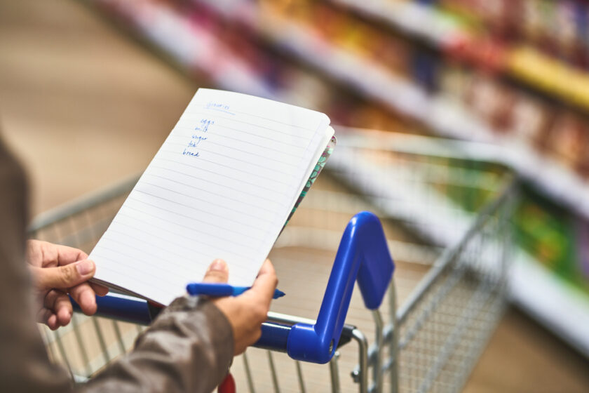 Hands, person and shopping list at grocery shop with wheel cart for foods, products and prices. Closeup, customer and supermarket or convenience store for retail cost, sale and discount or promotion.