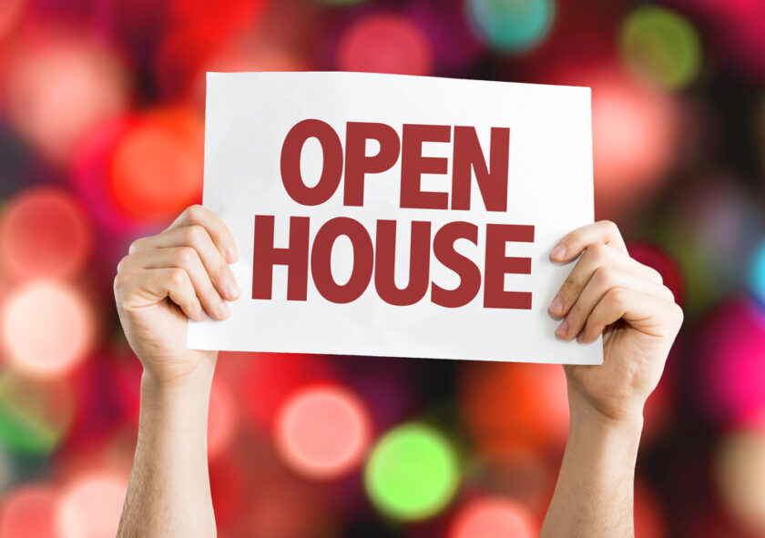 Two hands hold up a white sign with the words OPEN HOUSE in large red letters. The background is blurred with colorful bokeh lights.