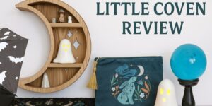 A crescent moon-shaped shelf holds small objects next to ghost figurines, a blue crystal ball, a pouch with a mystical design, and an umbrella with bat patterns. Text reads “LITTLE COVEN REVIEW.”.