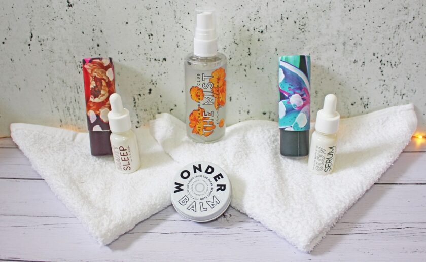 Skincare products, including bottles, tubes, and a tin labeled WONDER BALM, are arranged on a folded white towel against a light textured background. Two dropper bottles are labeled SLEEP and GLOW.