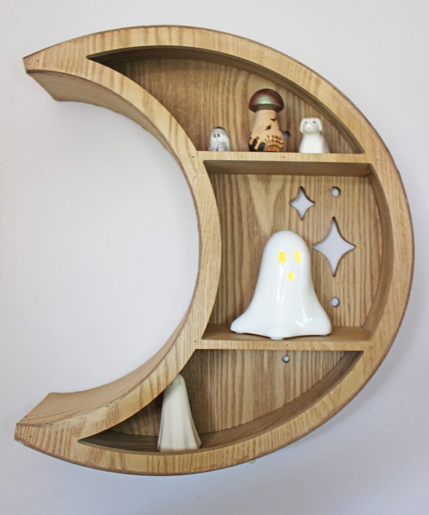 A wooden crescent moon-shaped wall shelf holds small ceramic figures, including ghosts and a person in a hat, against a plain white wall. The shelf has cutout star shapes on one compartment.