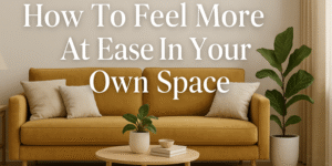 A cozy living room with a mustard yellow sofa, light cushions, a small round coffee table with plants and books, a large green potted plant, and beige curtains. Text reads: How To Feel More At Ease In Your Own Space.