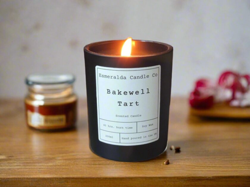 A lit candle in a dark glass jar labeled Bakewell Tart sits on a wooden surface. In the background, there’s a small jar and some blurred red and white objects. The scene is cozy and inviting.