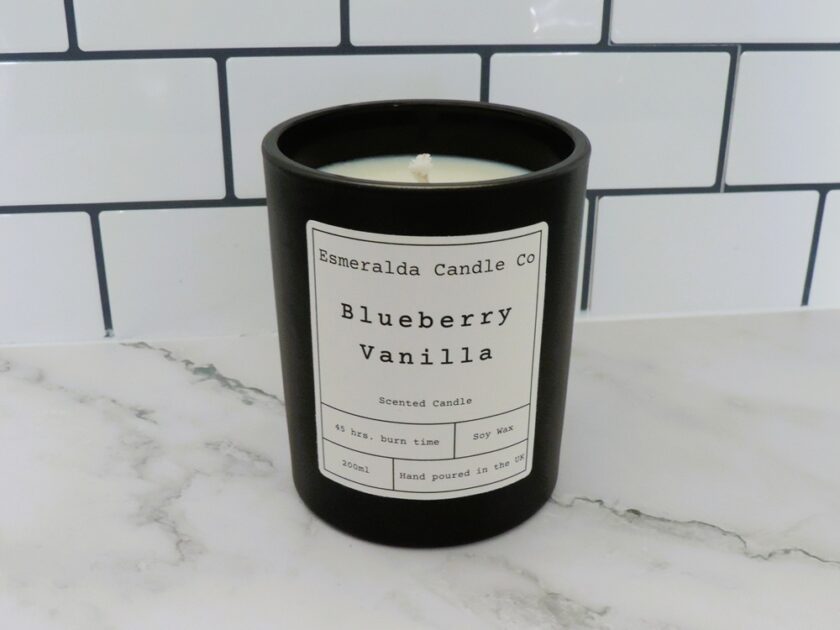 A black candle jar labeled “Esmeralda Candle Co Blueberry Vanilla” sits on a white marble counter in front of white subway tiles. The label details include 45 hr. burn time, soy wax, and 220ml, hand poured in the UK.
