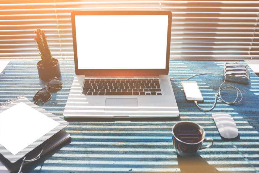 A laptop with a blank white screen sits on a blue striped desk, surrounded by a cup of coffee, smartphone, tablet, sunglasses, wallet, mouse, charging cable, and a small potted cactus. Sunlight streams through window blinds.