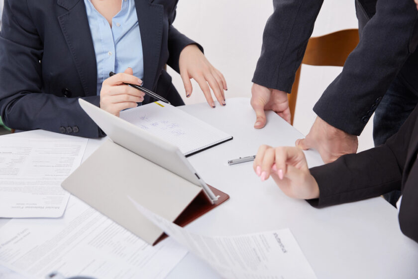 Three people in business attire sit and stand around a table with documents, a tablet, and a notebook, appearing to discuss paperwork or collaborate on a project. Only their hands and torsos are visible.
